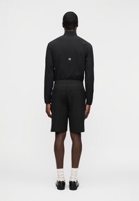 Άνδρας που στέκεται με την πλάτη γυρισμένη, φορώντας μαύρο μακρυμάνικο μπλουζάκι και σορτς, λευκές κάλτσες και μαύρα παπούτσια, μπροστά σε απλό φόντο.