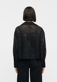 Γυναίκα με ίσιο καστανό καρέ κούρεμα, φοράει διαφανές μαύρο μπλουζάκι με floral μοτίβο και μακριά μανίκια και μαύρο παντελόνι, γυρισμένη πλάτη σε απλό φόντο.