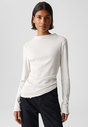 Camicia bianca a maniche lunghe con collo alto, texture morbida e orlo grezzo, abbinata a pantaloni neri. Design pulito e minimalista.