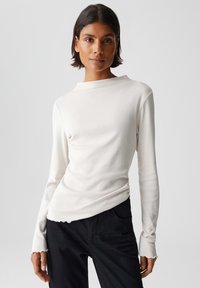 Weißes Langarmshirt mit hohem Kragen, weicher Textur und rohem Saum, kombiniert mit schwarzen Hosen. Sauberes und minimalistisches Design.