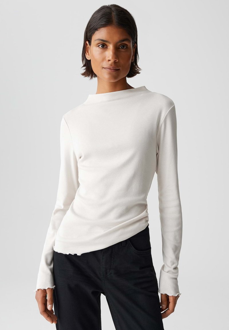 Weißes Langarmshirt mit hohem Kragen, weicher Textur und rohem Saum, kombiniert mit schwarzen Hosen. Sauberes und minimalistisches Design.