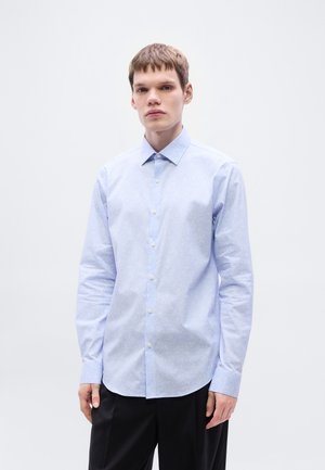 Jasnoniebieska koszula na guziki o subtelnym wzorzystym fakturze, z długimi rękawami i zaostrzonym kołnierzem, zestawiona z ciemnymi spodniami.