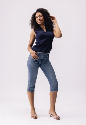 Marine blauw mouwloos poloshirt gecombineerd met lichtblauwe cropped jeans. De outfit heeft een geribbelde textuur en een klassieke kraag. Het model draagt doorzichtige hakken.