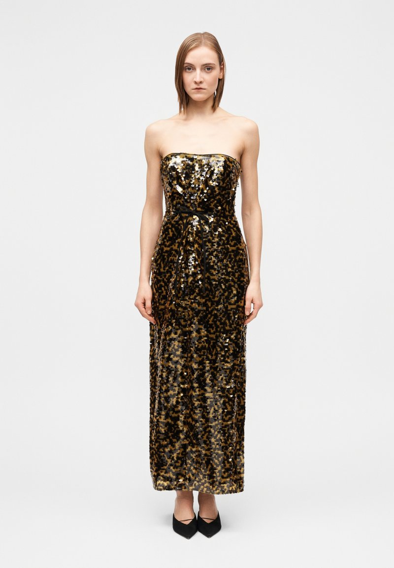 Woman wearing a strapless, floor-length sequined dress with black and gold pattern, paired with black pointed shoes, standing on a white background.