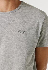 Nærbillede af en person iført en lysegrå T-shirt med "Pepe Jeans London"-logo på venstre bryst.