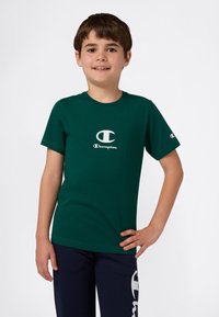 Ragazzo sorridente che indossa una maglietta verde Champion e pantaloni della tuta blu navy con il logo Champion, in piedi con una mano sui fianchi.