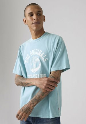 Mężczyzna z tatuażami w jasnoniebieskiej koszulce "The Originals Levi Strauss", z założonymi na piersi ramionami, patrzący w bok na jednolitym tle.