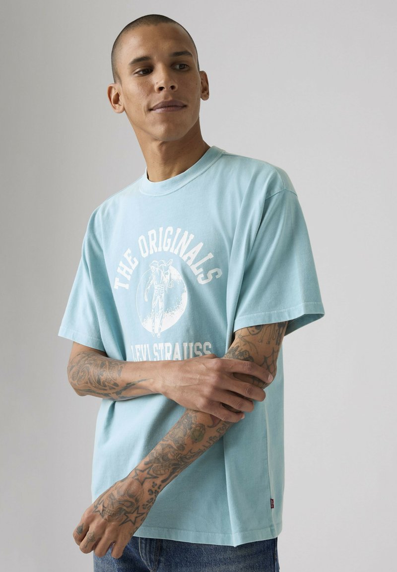 Hombre con tatuajes que lleva una camiseta azul claro con el texto "The Originals Levi Strauss", cruzando los brazos y mirando hacia un lado sobre un fondo liso.