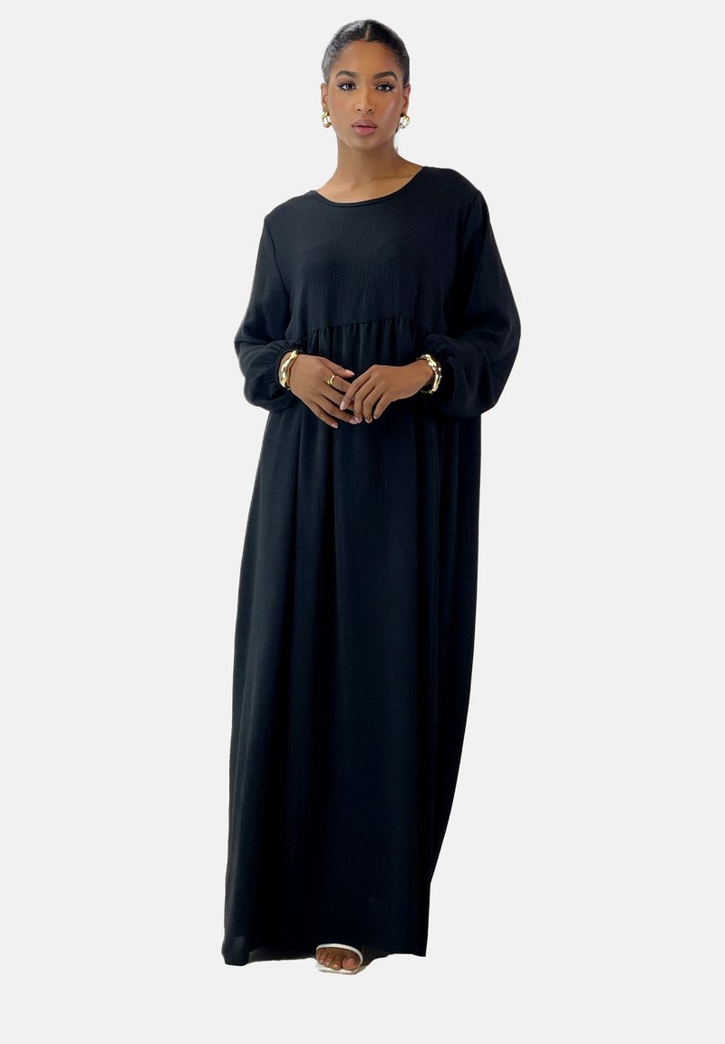 Vestido maxi negro con mangas largas, tejido texturizado, corte suelto y cintura en capas. Accesorios de joyería dorada en los brazos del modelo.