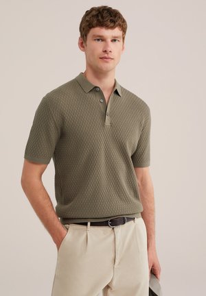 Junger Mann trägt ein strukturiertes olivgrünes kurzärmeliges Poloshirt mit beigen Hosen und schwarzem Gürtel, steht mit einer Hand in der Tasche.
