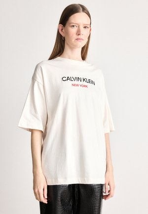 Heledbeez värvi oversized T-särk, millel on musta ja punase tikandiga tekst "CALVIN KLEIN NEW YORK". Mudel kannab musti tekstuuriga pükse.