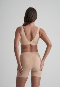 Kvinna sett bakifrån iförd beiga sömlösa högmidjade shorts och en matchande behå mot en enfärgad ljus bakgrund.