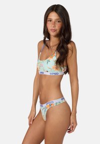Femme aux longs cheveux foncés portant un ensemble soutien-gorge et culotte imprimé floral avec des bandes élastiques blanches étiquetées PSD, debout devant un fond uni.