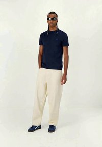 Bărbat îmbrăcat cu ochelari de soare negri, tricou albastru închis, pantaloni largi crem și adidași navy cu dungi albastre deschise.