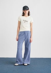 Νεαρή γυναίκα φορεί καπέλο Nike μαύρου χρώματος, κρεμ μπλούζα Nike, μπλε φαρδιά παντελόνια φόρμας και λευκά αθλητικά παπούτσια, ενώ στέκεται πάνω σε μπλε χαλί.