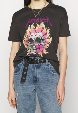 Persoon draagt een zwart Metallica T-shirt met brandende schedel en roze bloemmotief, blauwe spijkerbroek, zwarte riem en gelaagde gouden kettingen.