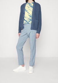 Blå stickad kofta med knappar, t-shirt med gul och blå batikmönster, ljusblå skräddarsydda byxor och vita sneakers.