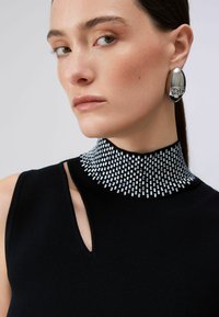 Femme aux cheveux lisses portant un haut noir sans manches avec des ornements rectangulaires argentés sur le col haut et une grande boucle d'oreille ovale argentée.