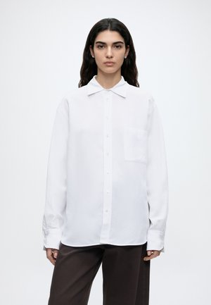 Młoda kobieta o długich ciemnych włosach, ubrana w luźną białą koszulę na guziki i ciemne spodnie, stojąca na jednolitym białym tle.