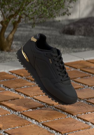 Black athletic shoe with a textured surface, gold accent on the heel, and a rugged rubber sole, suspended over a patterned stone path.