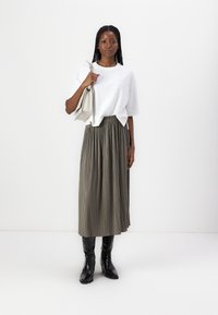 Tricou alb cu mânecă scurtă și detalii texturate, asortat cu o fustă plisată de culoare verde măsliniu și cizme negre până la genunchi; ținând o geantă de culoare gri deschis.