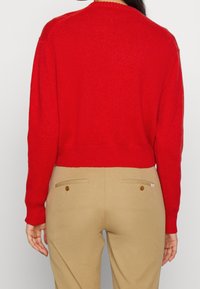 Henkilö, joka on pukeutunut kirkkaan punaisiin pitkähihasiin neuleisiin ja beigearvoisiin housuihin, joissa on napitetut takataskut, näkyy vyötäröstä ylös, takaa päin.