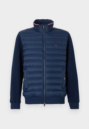 Marineblauw donzen jack met een hoge kraag, geribbelde manchetten, ritssluiting en horizontale doorgestikte secties. Voorzien van contrasterende biezen en zijzakken.