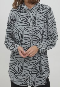 Camisa de botones en gris claro con un patrón de rayas de cebra negras. Cuenta con mangas largas, un collar y un ajuste relajado con un dobladillo curvado.