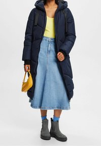 Langer navyblauer Puffer-Mantel, gelbes Tank-Top, Jeans-A-Linien-Rock, graue Ankle-Boots und eine kleine gelbe Handtasche. Mit schwarzen Akzenten und Details.