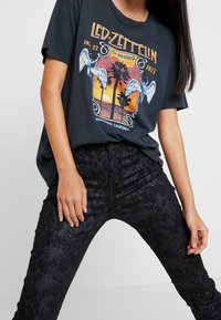 Černé grafické tričko s designem Led Zeppelin, s živými barvami západu slunce a palmami. V kombinaci s černými kalhotami s hadím vzorem.