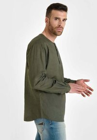 Olijf groene blouse met lange mouwen en een ronde halslijn, zachte textuur en knoopsluiting. Gecombineerd met lichtblauwe denim jeans.