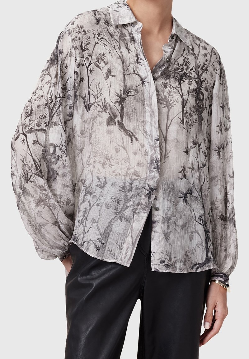 Blouse grise semi-transparente à motifs floraux et oiseaux noirs, manches longues bouffantes, devant boutonné, associée à un pantalon en cuir noir.