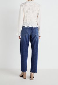 Bílá dlouhá halenka s nařaseným detailem na zadní straně, v kombinaci s modrými vysokými džínami, které mají rovný střih a dvě zadní kapsy.