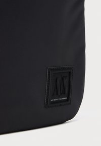 Borsa in tessuto nera con una toppa quadrata cucita che mostra il testo in rilievo "A|X" e "Armani Exchange" nell'angolo in basso a destra.