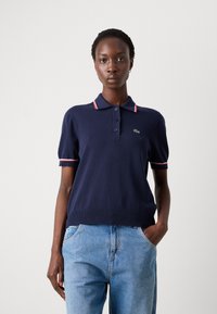 Tamsiai mėlyna trumpomis rankovėmis polo marškinėliai su raštuotu apykakle, papuošta raudonomis ir baltomis detalėmis. Mygtukai priekyje ir Lacoste logotipas ant krūtinės.