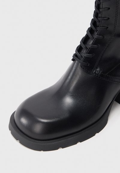 Bota de cuero negro para tobillo con punta redondeada, acabado texturizado, cordones delanteros y suela gruesa de goma negra con detalles acanalados.