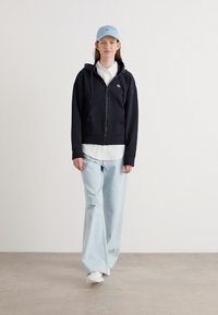 Sudadera con cremallera azul marino sobre una camisa blanca con cuello, acompañada de jeans de pierna ancha en azul claro y zapatillas blancas. Una gorra azul clara completa el conjunto.