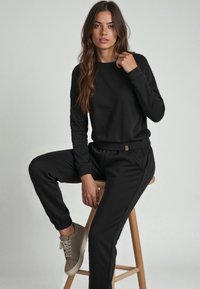 Svart sweatshirt med långa ärmar och rund halsringning, parat med matchande joggerbyxor med elastiska muddar och en liten varumärkesetikett. Beige sneakers.