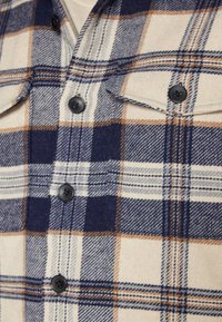 Zoom su una camicia a scacchi crema, blu navy e beige con bottoni neri e due tasche sul petto con bottone.