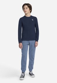 Chemise à manches longues bleu marine avec un petit logo blanc, associée à un pantalon de jogging bleu clair et des baskets noires avec des accents blancs. Design simple dans l'ensemble.