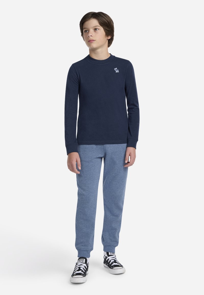 Chemise à manches longues bleu marine avec un petit logo blanc, associée à un pantalon de jogging bleu clair et des baskets noires avec des accents blancs. Design simple dans l'ensemble.