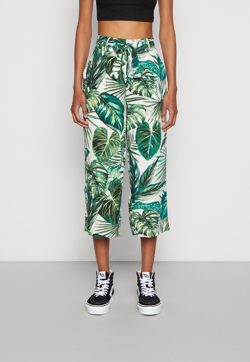 Spodnie culotte z zielonym wzorem liści na białym tle, z wiązanym pasem i luźnym krojem. Połączone z czarnymi trampkami za kostkę.