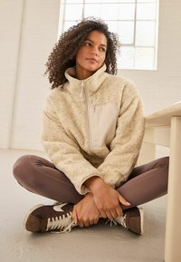 Fleece-Zipperjacke in Creme mit strukturierter Oberfläche, ausgestattet mit einer beigen Brusttasche und hohem Kragen. Tragen mit braunen Leggings und Sneakers.