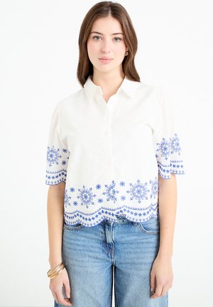 Femme portant une chemise blanche à manches courtes avec une broderie florale bleue, associée à un jean bleu clair et des bracelets dorés au poignet gauche.