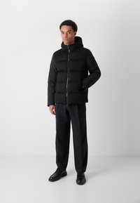 Black insulated jacket with a zip front, hood, and quilted pattern on the chest. Worn with dark trousers and black shoes. Minimalist design.