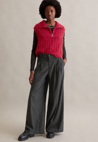 Rotes gestricktes ärmelloses Oberteil mit einem Reißverschluss am Hals, kombiniert mit hochgeschnittenen, dunkelgrauen, weit geschnittenen Hosen und schwarzen Schuhen mit dicker Sohle.