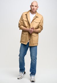Chaqueta de algodón color beige con cuatro bolsillos delanteros, llevada sobre una camiseta blanca, combinada con vaqueros azules y zapatillas grises. Diseño simple y estilo práctico.