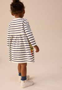 Vestito a maniche lunghe a righe in avorio e blu navy con vita arricciata, decorato con un'applicazione colorata a forma di faccina sorridente di lato. Sneakers bianche con calzini blu.