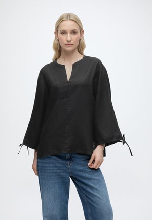 Žena s dlhými blond vlasmi má na sebe čiernu voľnú blúzku so širokými rukávmi a modré džínsy, stojaca pred jednoliatym bielym pozadím.