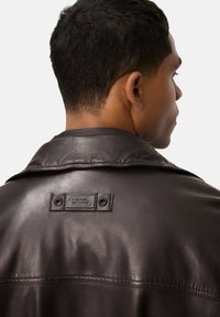 Bruine leren jas met een brede kraag, voorzien van een rechthoekig merklabel en twee drukknoopsluitingen aan de achterkant. Gladde textuur en getailleerde snit.